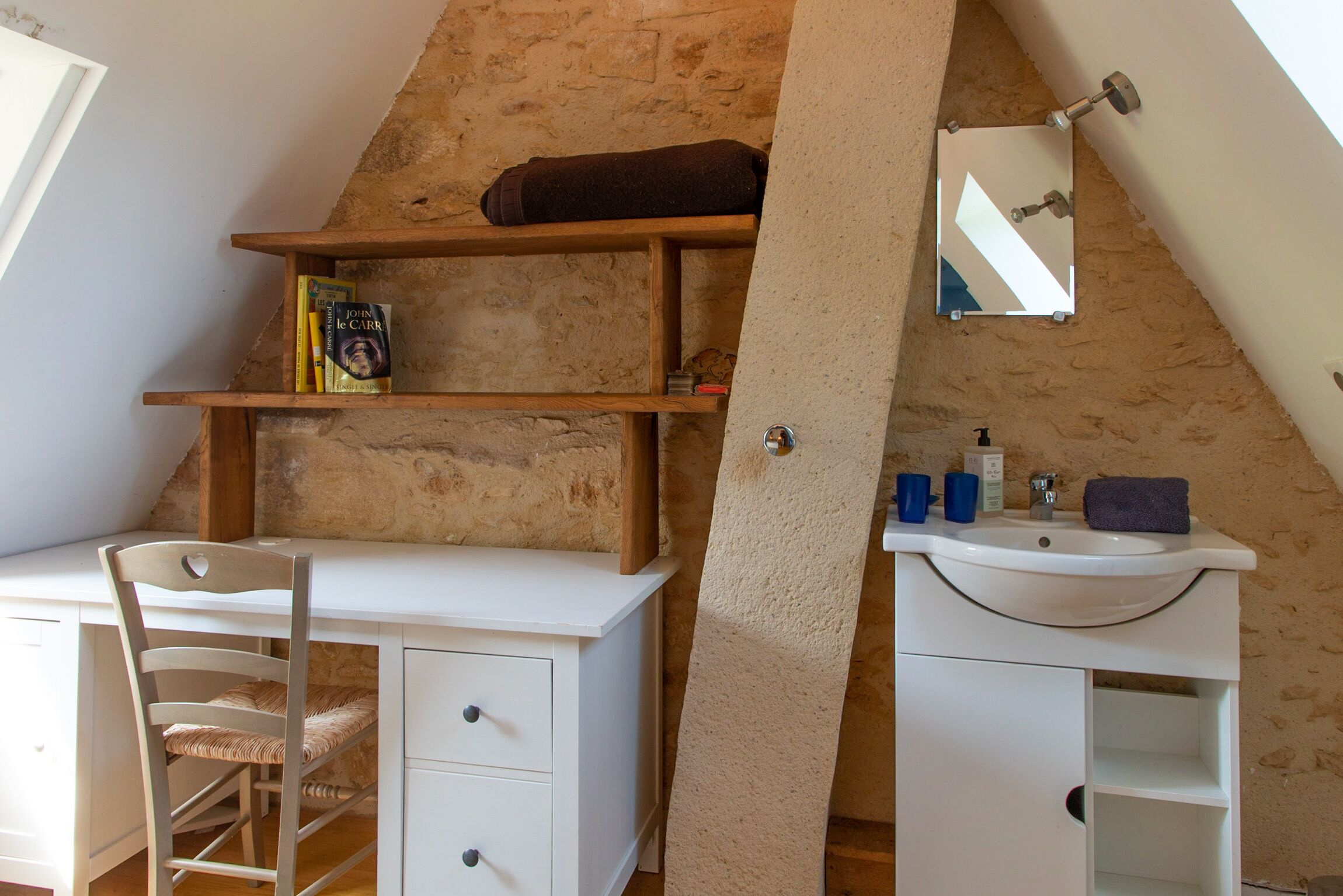 Ein Schlafzimmer mit schrägverdeckter Decke, einem weißen Schreibtisch mit Stuhl, einem Bücherregal, einem Spiegel und einem Badezimmer mit Waschbecken und Handtuch.