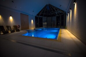 A swimming pool with a glass ceiling, lounge chairs, and wall-mounted lights.