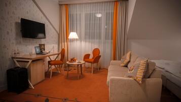 Habitación de hotel con escritorio, dos sillas naranjas, un sofá con cojines estampados, un televisor montado en la pared y una ventana con cortinas traslúcidas.