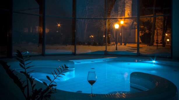 A glass-enclosed hot tub with a glass of red wine, surrounded by a snowy landscape and illuminated by street lamps.