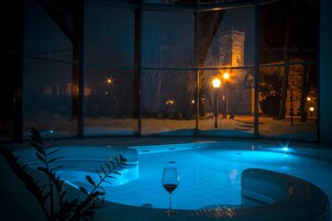 Un jacuzzi con paredes de vidrio, una copa de vino tinto, un paisaje nevado y farolas que lo iluminan.