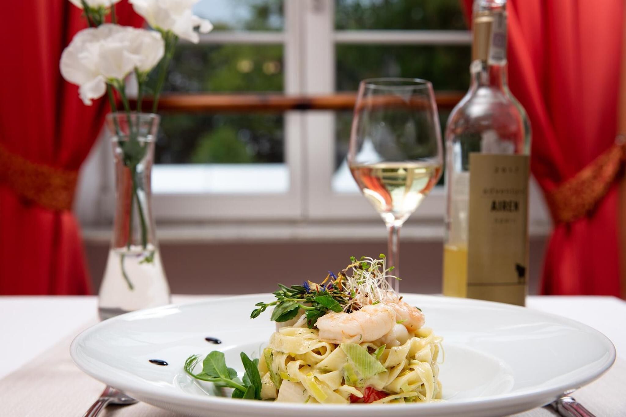 A plate of pasta with shrimp and greens, a wine glass, and a bottle of wine on a table with a window view.