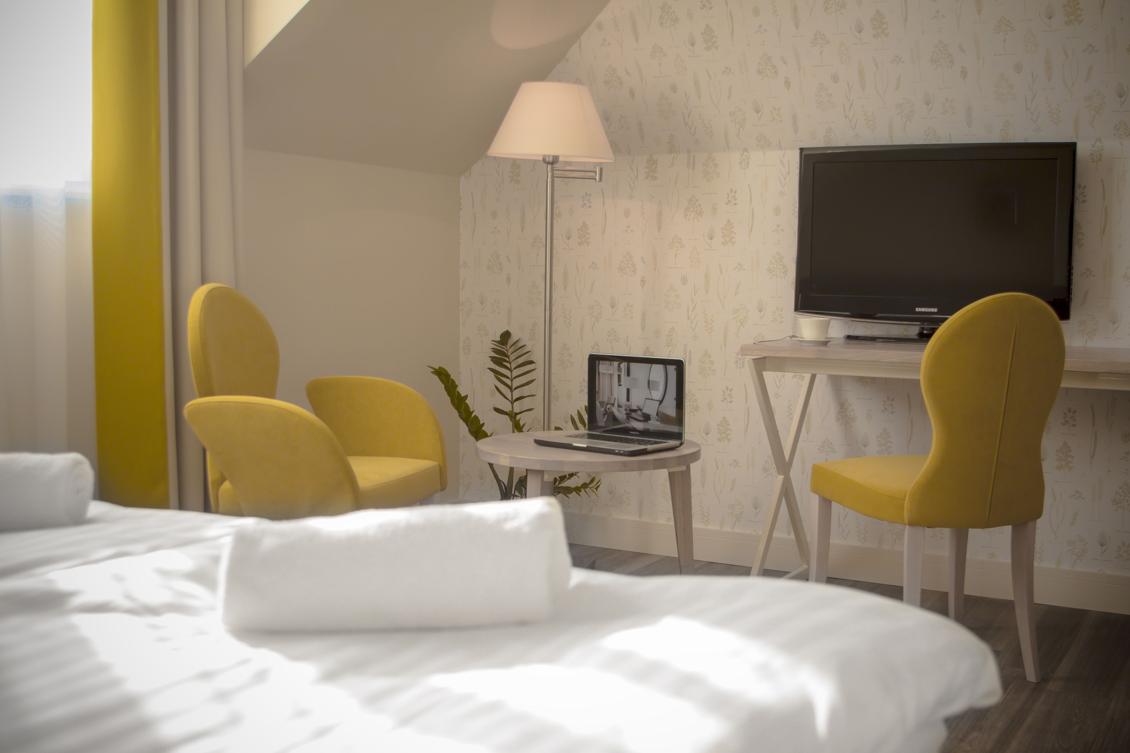 Habitación de hotel con una cama, dos sillones amarillos, un escritorio con una computadora portátil, un televisor y una lámpara de pie.