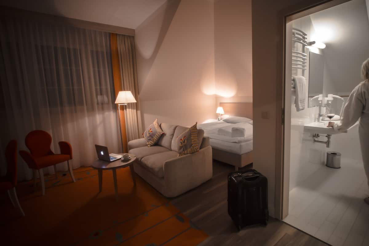 Une chambre d’hôtel avec un canapé, un fauteuil, un bureau avec un ordinateur portable, un lit, une valise et une salle de bain visible à travers une porte ouverte.