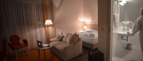 A hotel room with a sofa, a chair, a desk with a laptop, a bed, a suitcase, and a bathroom visible through an open door.