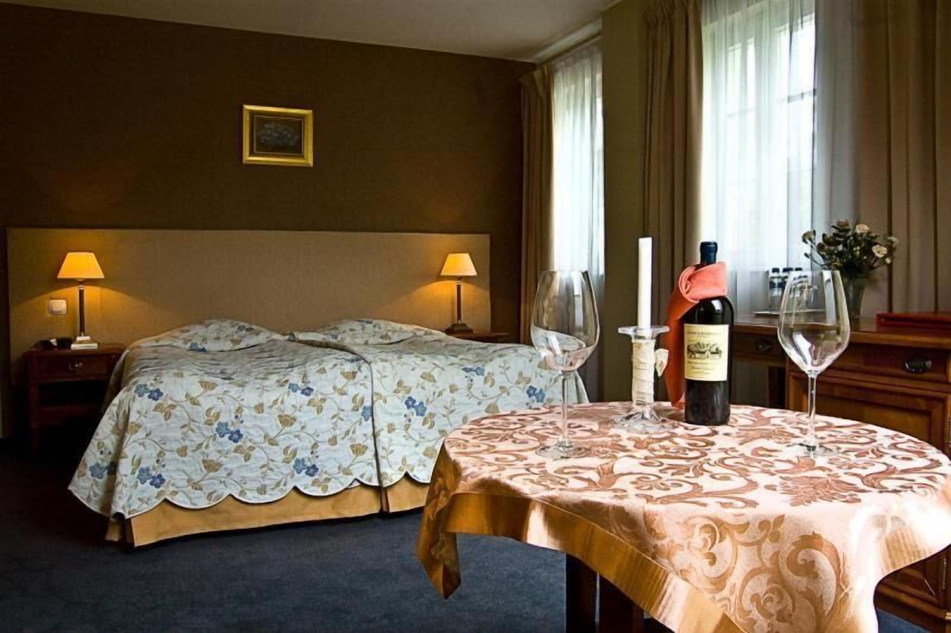 Une chambre d’hôtel avec un lit double, deux lampes de chevet, une table avec une bouteille de vin et un verre, et un vase rempli de fleurs.
