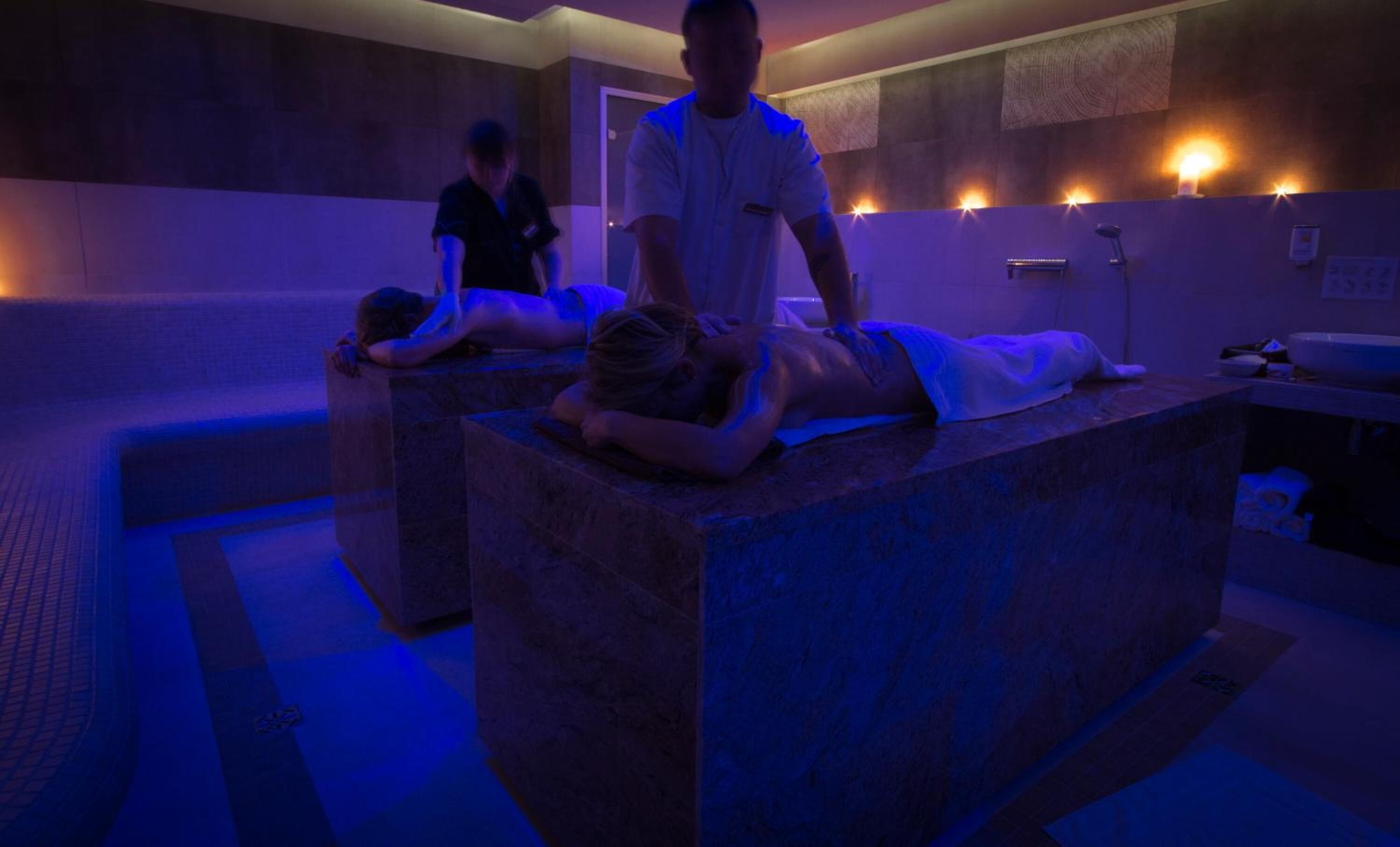 A massage therapy room with two massage tables and a therapist attending to clients.