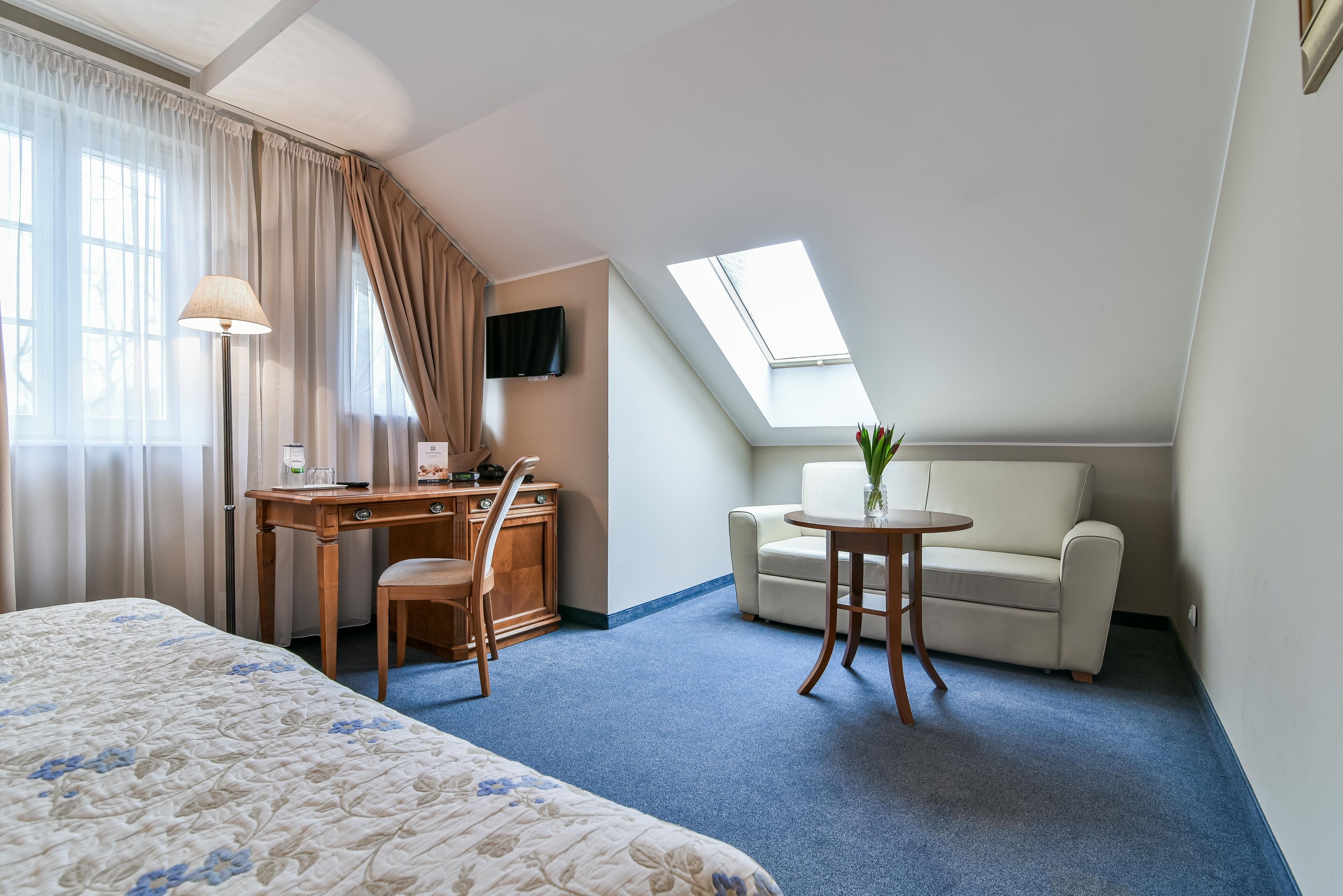 A hotel room with a bed, a desk with a chair, a small table with a vase, a sofa, and a TV on the wall.