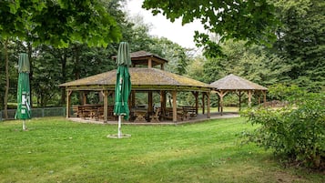 A gazebo with a tiled roof and wooden benches, surrounded by lush greenery and trees.