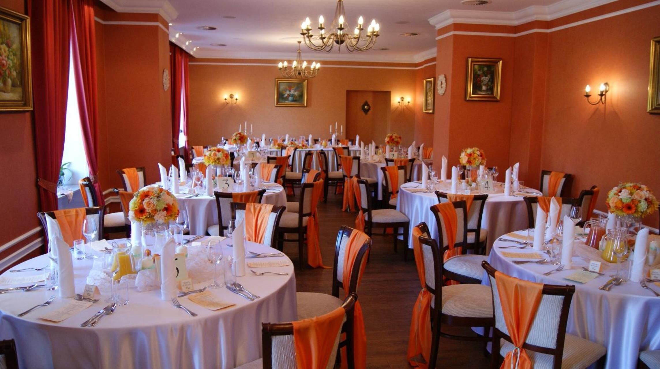 A banquet hall with round tables, orange chair sashes, and floral centerpieces.