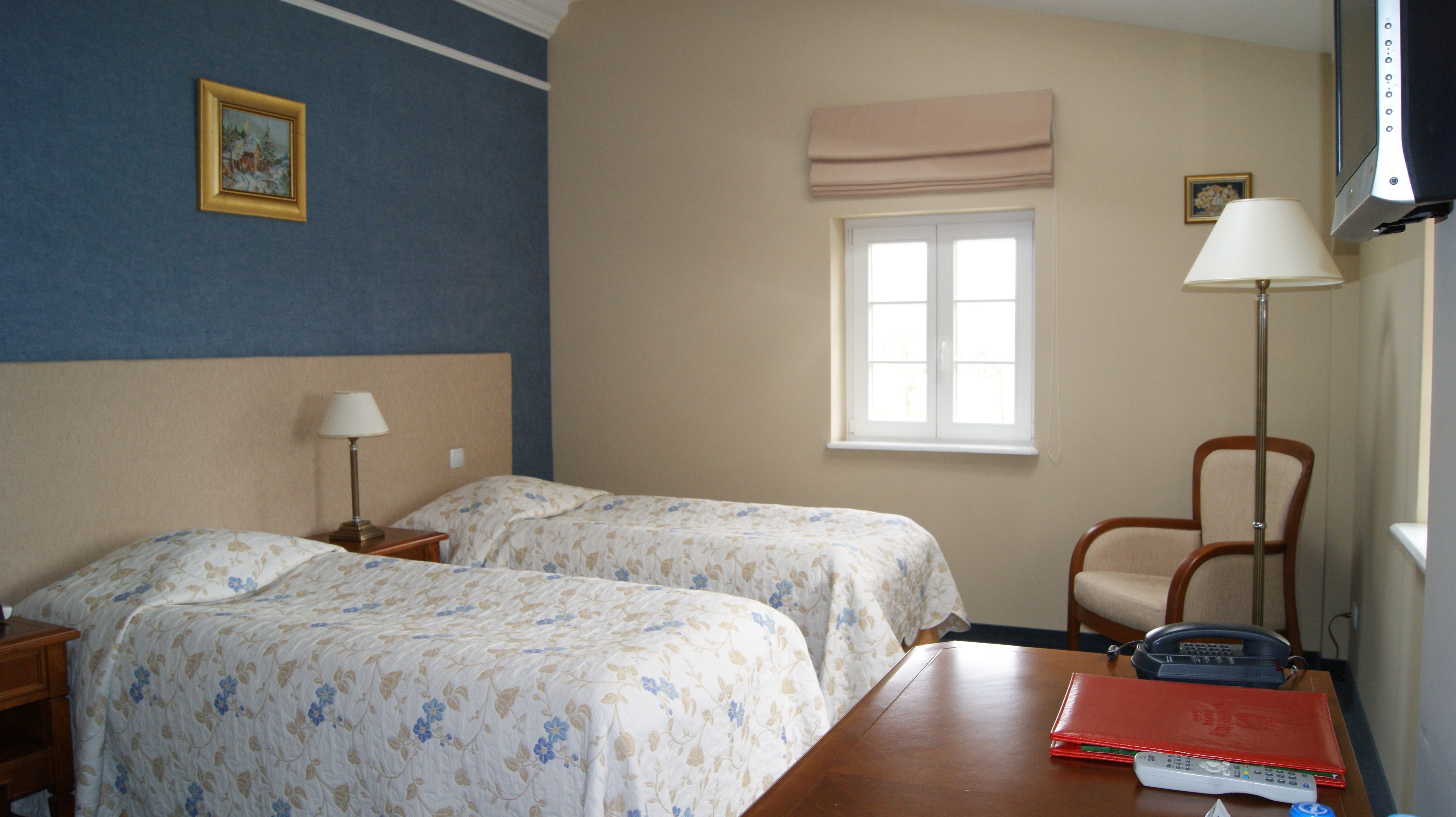 A hotel room with two beds, a window with a view, a chair, a desk with a lamp, and a TV.