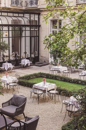 Un área de comedor al aire libre con mesas y sillas, un jardín y la fachada de un edificio con ventanas y balcones.