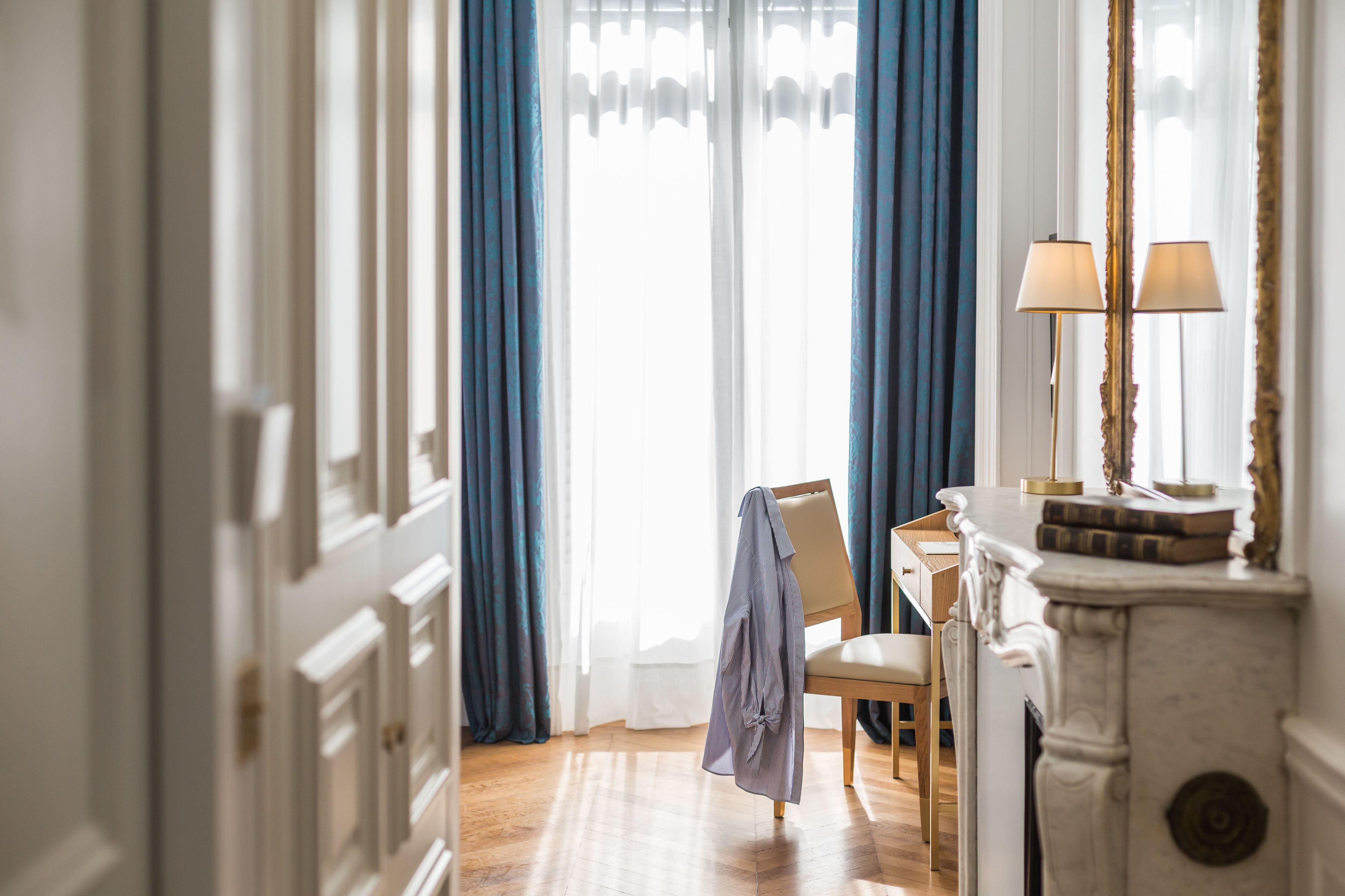 A room with a wooden floor, a chair, a table, a mirror with a lamp, and a window with blue curtains.