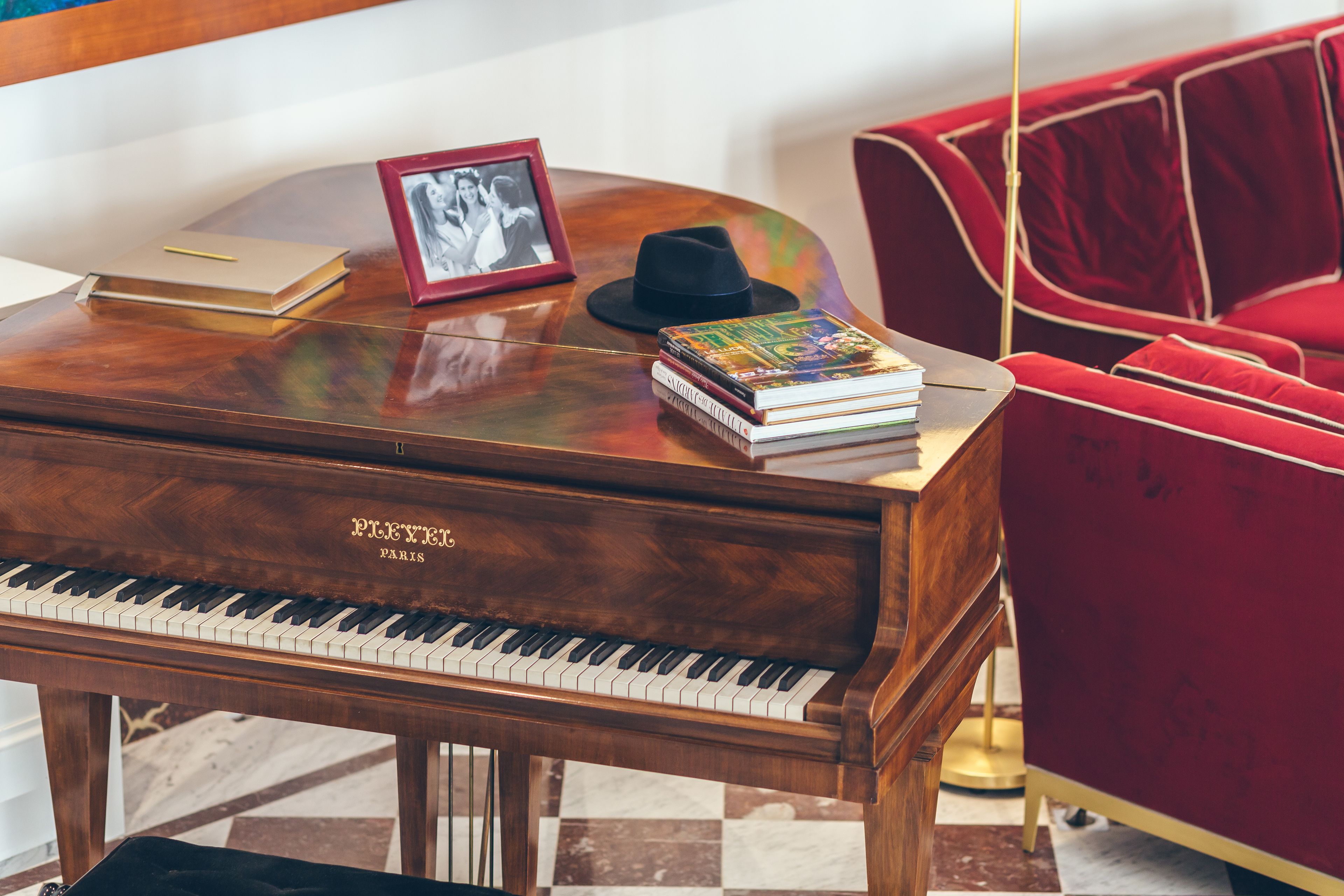 A grand piano, a red velvet armchair, a stack of books, a black hat, and a framed photo on a wooden table.