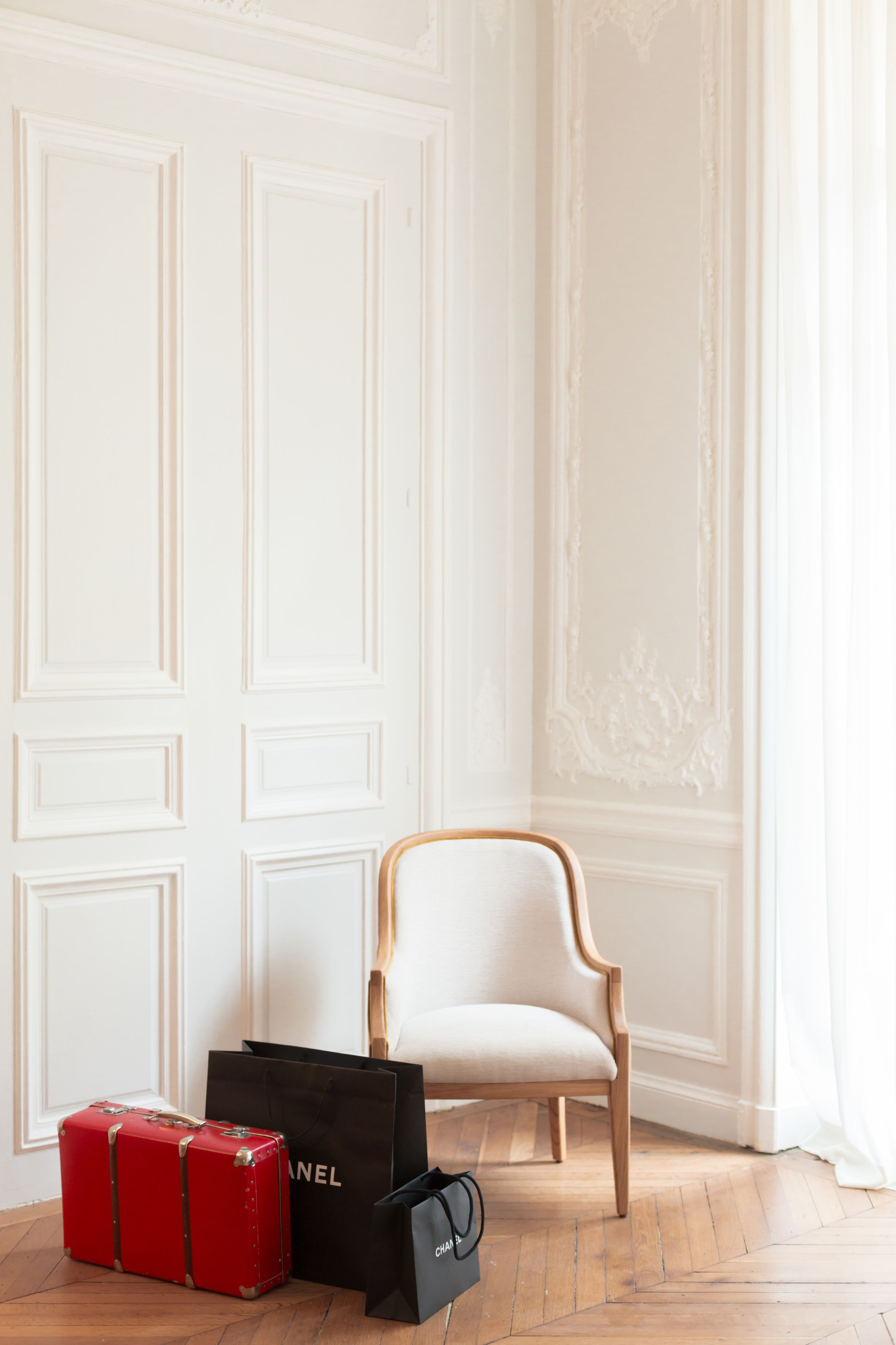 A white upholstered chair, a red suitcase, and two black shopping bags with the word "CHANEL" on them, placed on a wooden floor against a white wall with paneling.