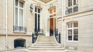 Une entrée imposante, avec un escalier menant à une porte, flanquée de balustrades en fer forgé richement décorées et de grandes fenêtres équipées de volets.