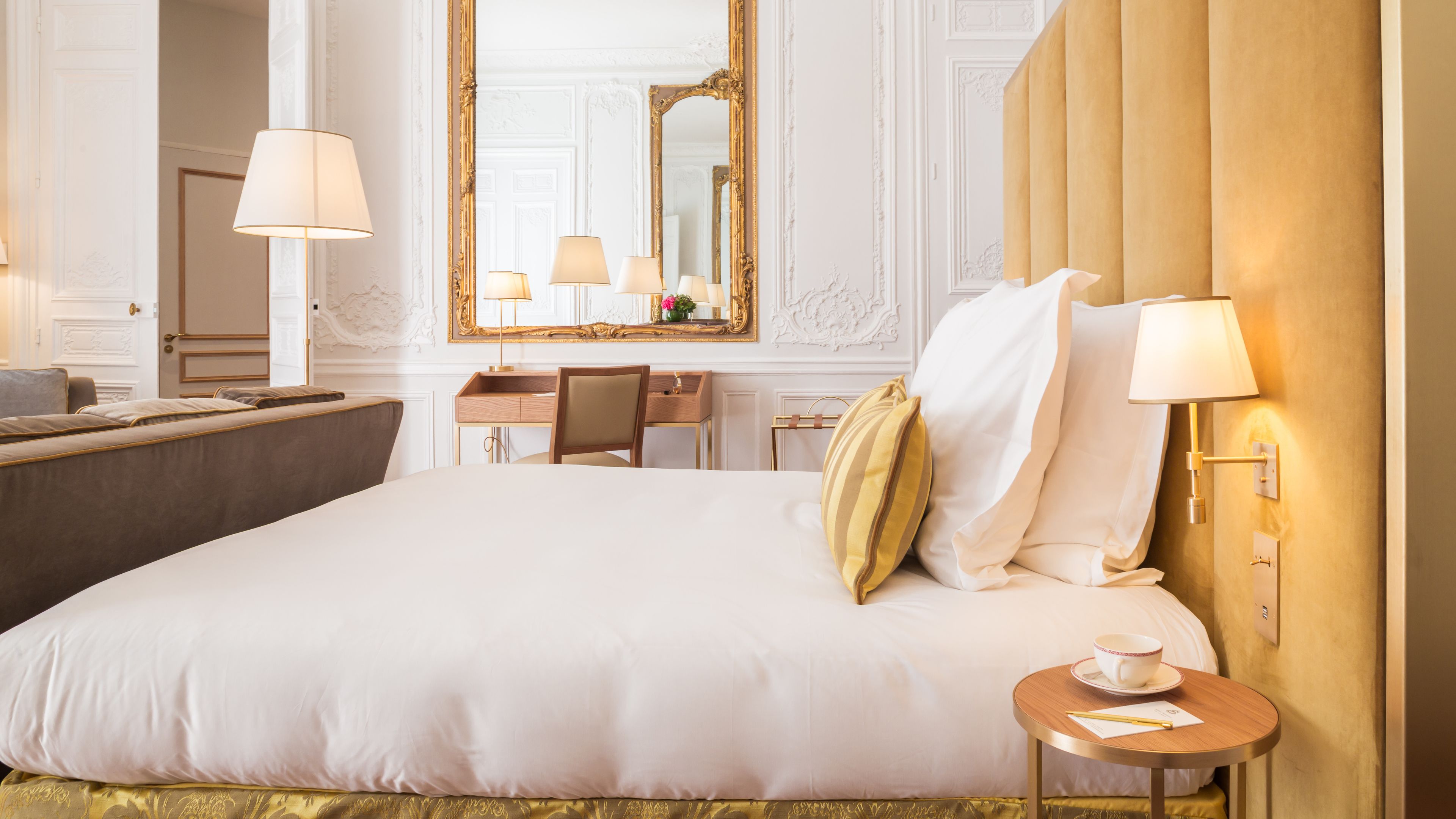 A hotel room with a large bed, a desk, a chair, a small table with a cup and a book, and a mirror with a gold frame.