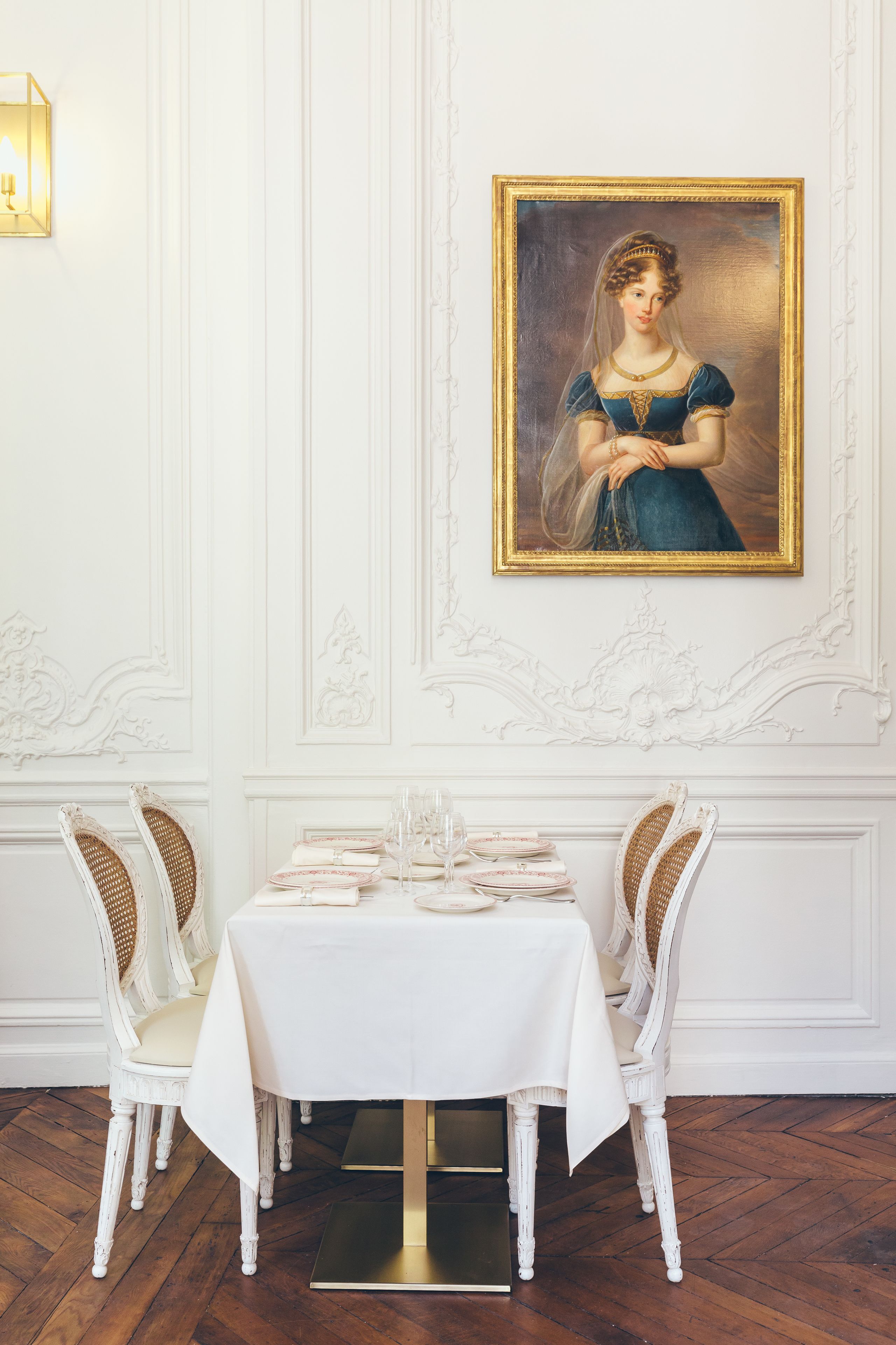 A dining room with a white table set for four, white upholstered chairs, and a framed portrait on the wall.