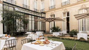 An outdoor dining area with tables set for a meal, surrounded by a garden and a building with ornate balconies. - Hotel Alfred Sommier (Paris)