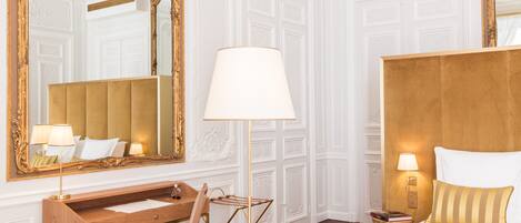 A hotel room with a wooden desk, a large mirror, a bed with a gold headboard, and a nightstand with a lamp.