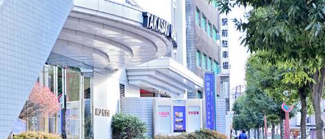 A modern building with a curved facade, signage for 'TAKASAKI', and a pedestrian walkway with trees and a person walking.