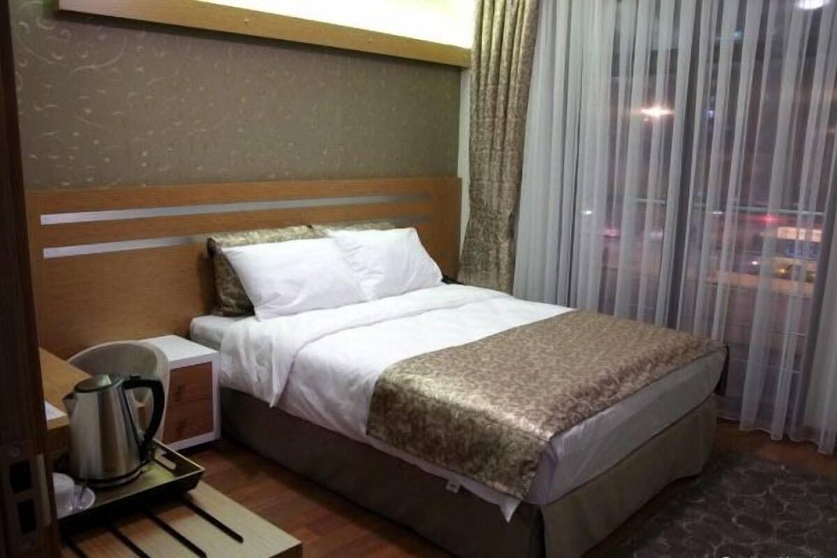 A hotel room with a bed, a nightstand, a kettle, and a view of the city through sheer curtains.