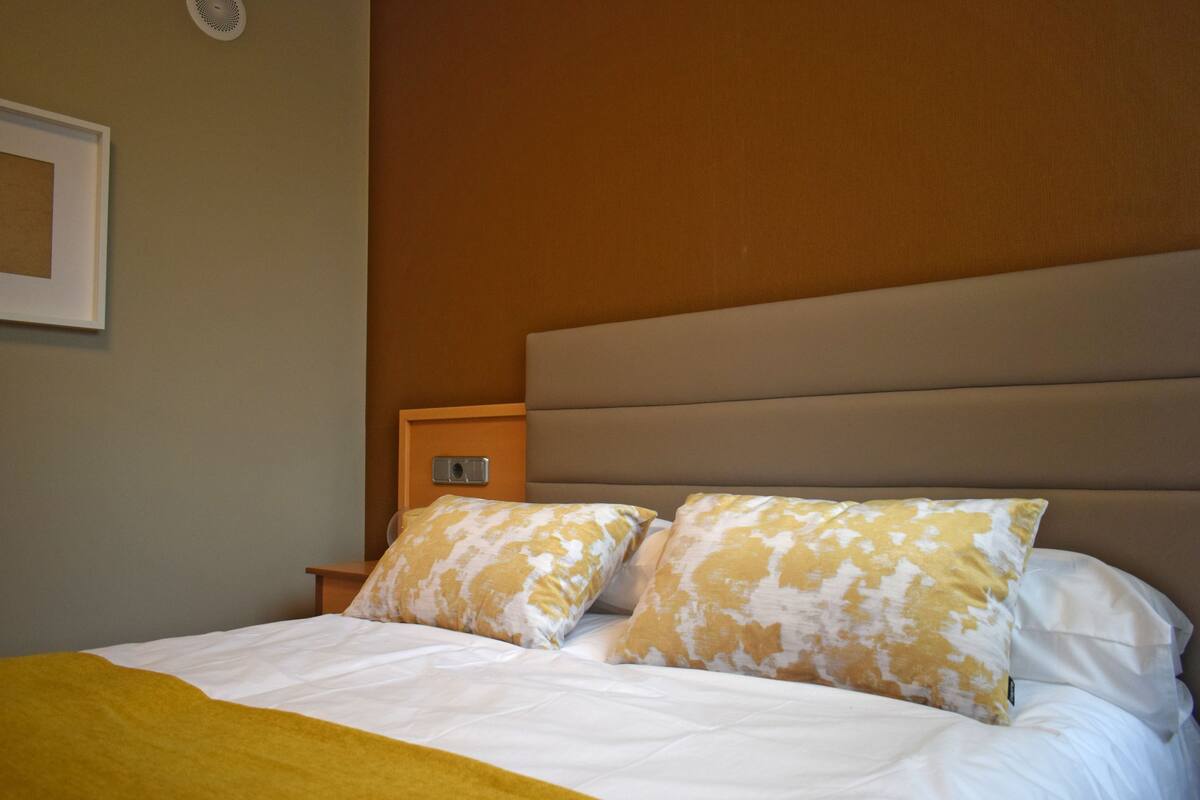 A neatly made bed with white and yellow patterned pillows, a wooden nightstand, and a beige headboard.