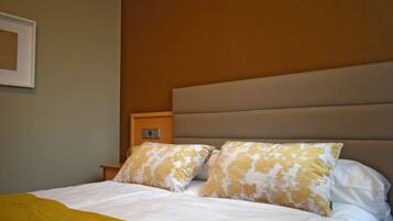 Una cama bien hecha con almohadas estampadas en blanco y amarillo, una mesita de noche de madera y un cabecero color beige.