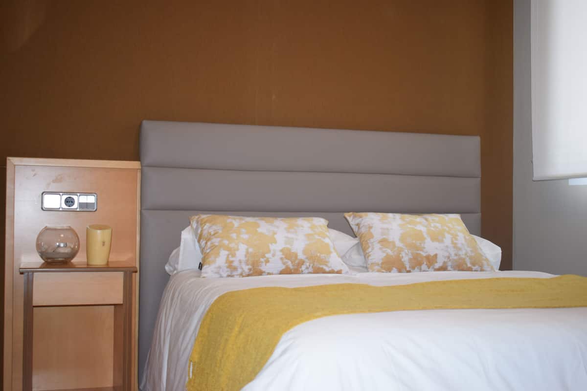 A neatly made bed with a white and yellow patterned pillow, a yellow throw blanket, and a wooden nightstand with a vase and a lamp.