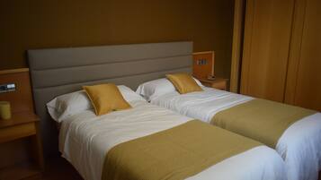 A double bed with white bedding and two mustard-colored pillows, a wooden nightstand with a lamp, and a wooden wardrobe.