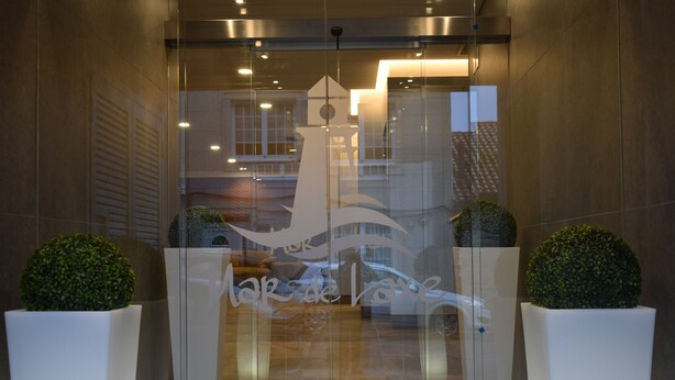 Entrada moderna de un hotel con puerta de cristal, dos plantas en macetas y el reflejo de un edificio y un coche.