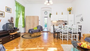 A compact living space with a wooden floor, a sofa, a dining table with chairs, a kitchen area, and a small TV on a wooden cabinet.