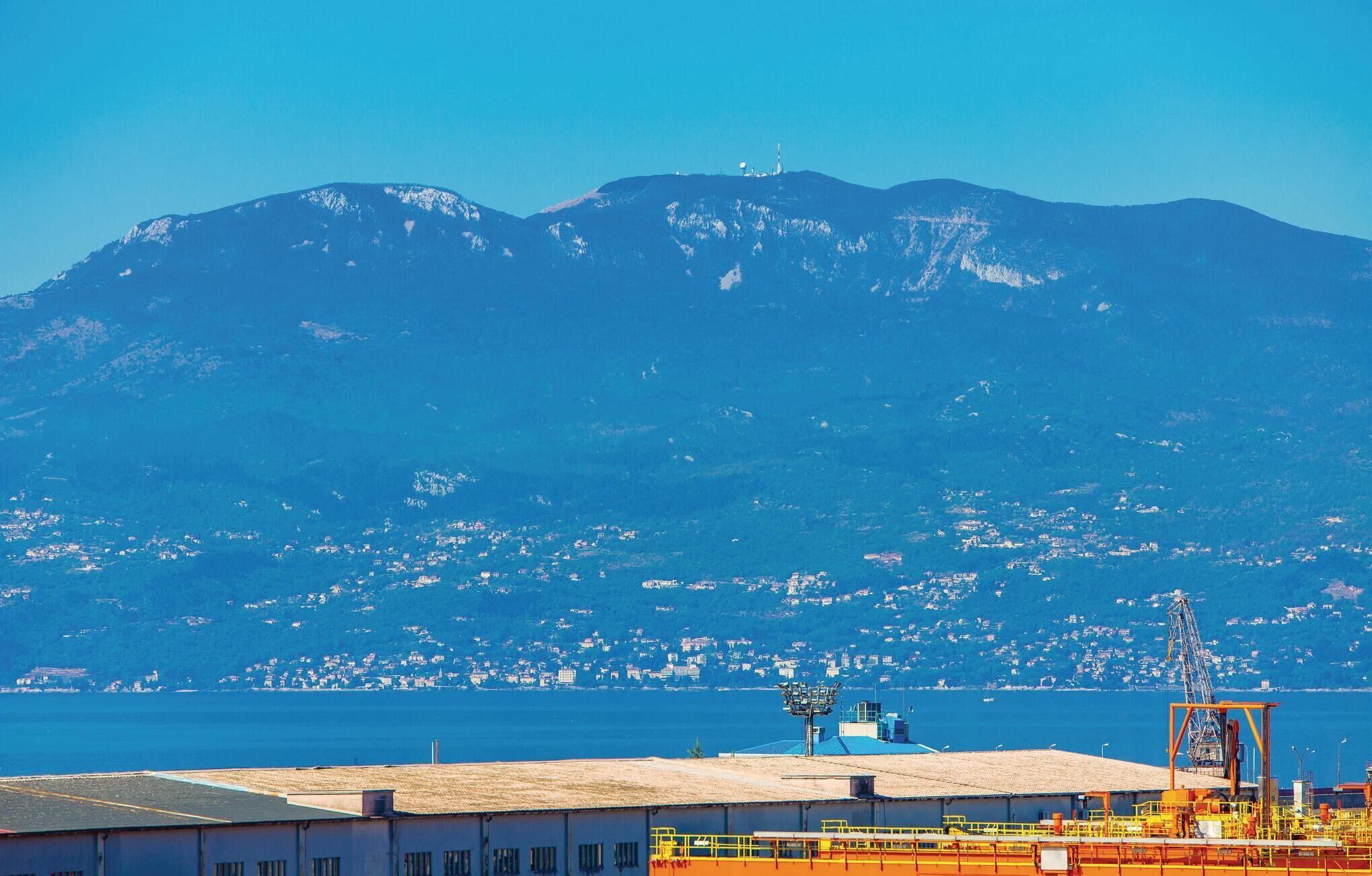 A coastal view with a mountain in the background, a harbor with industrial structures, and a small town across the water.