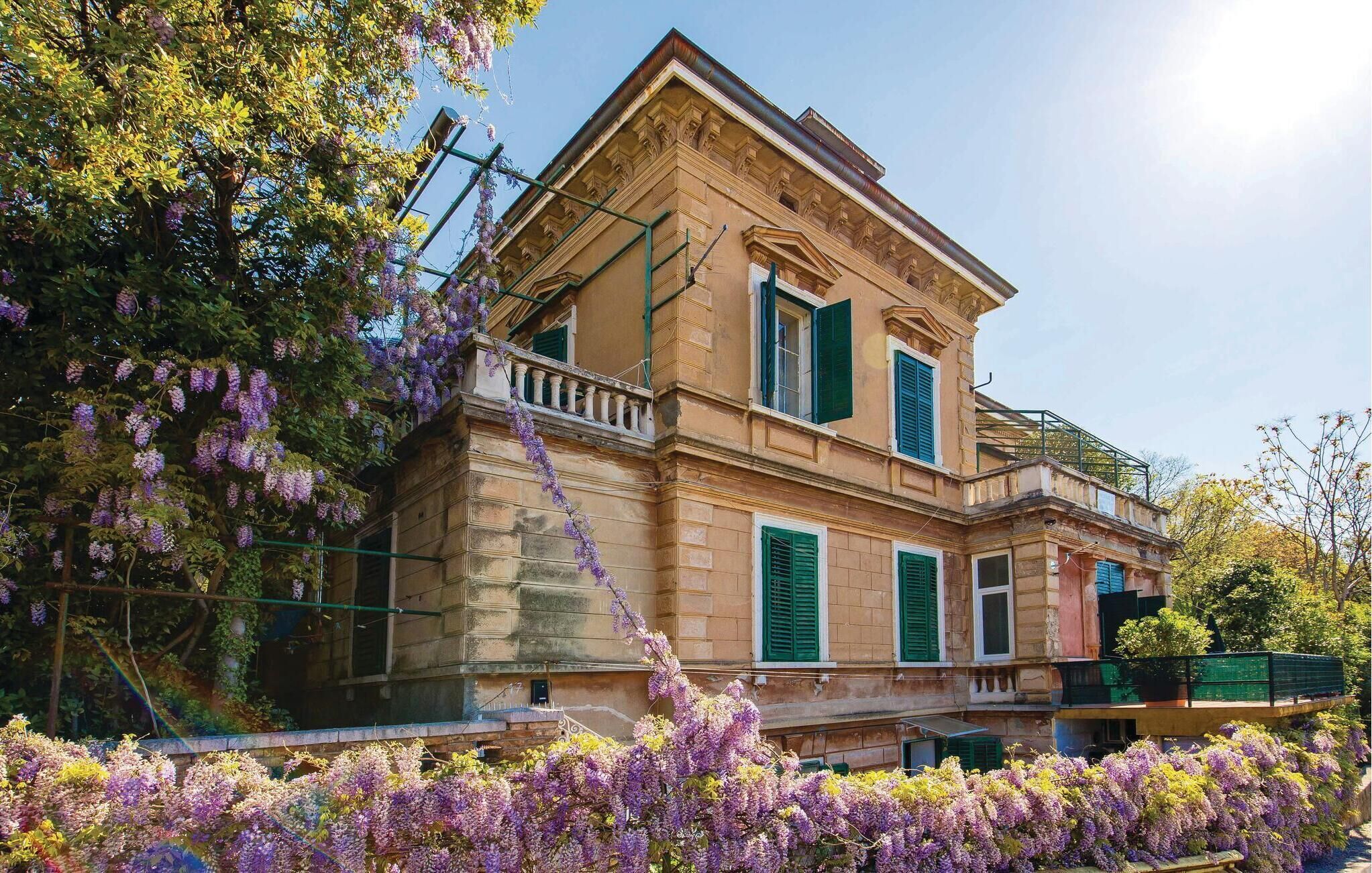 Ein zweistöckiges Gebäude mit Balkon, grünen Fensterläden und einem Garten mit violetten Blumen.