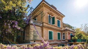 A two-story building with a balcony, green shutters, and a garden with purple flowers.