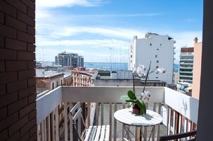 Un balcón con mesa y sillas blancas, una planta en maceta y vistas al mar y edificios.