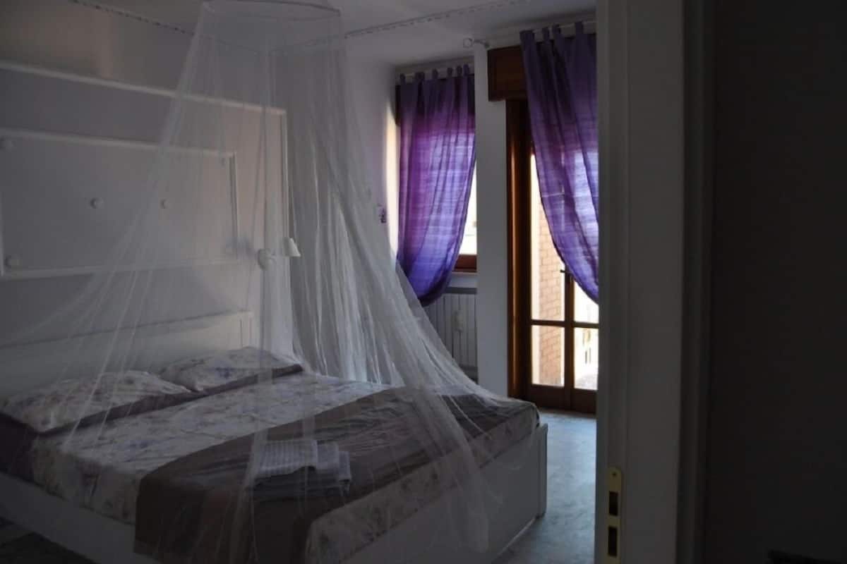 Une chambre à coucher avec un lit recouvert d’une moustiquaire, des rideaux violets et une vue sur l’extérieur.