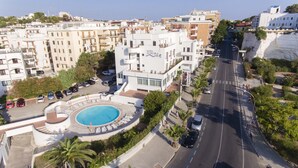 Un hotel con piscina, zona de estacionamiento y una calle con coches y peatones.