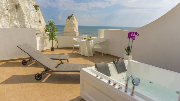 Una terraza en la azotea con una tumbona, una mesa con sillas, una planta pequeña en maceta y una bañera con vistas al mar y los acantilados.