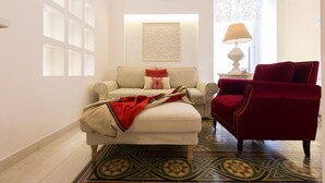 A living room with a beige sofa, a red armchair, patterned floor tiles, a decorative wall piece, and a window with a curtain. - Suite Le Perle (Taranto)