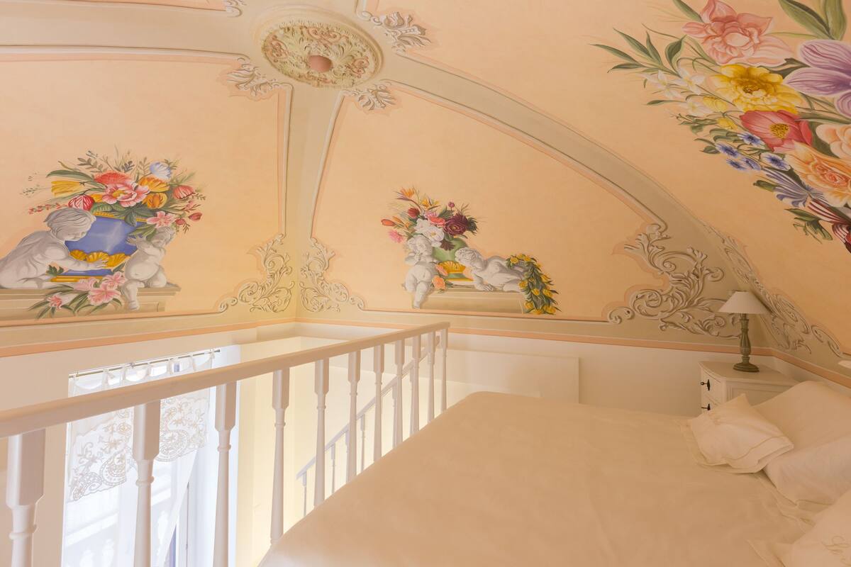Ein Schlafzimmer mit einem weißen Bett, einem Nachttisch, einer Lampe und einer dekorativen Decke mit floralen und Engelsmotiven.