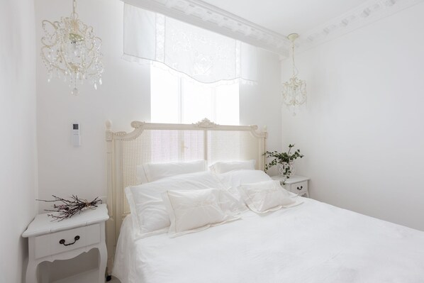 A white bedroom with a bed, two nightstands, a window with sheer curtains, and two chandeliers.