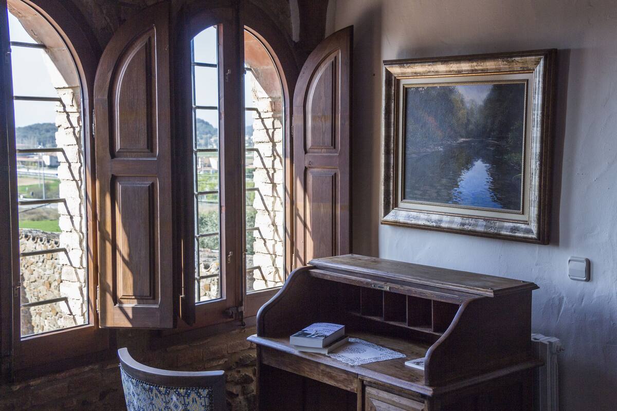 Une pièce comprenant un bureau, un tableau encadré et trois fenêtres en arc donnant sur un paysage.