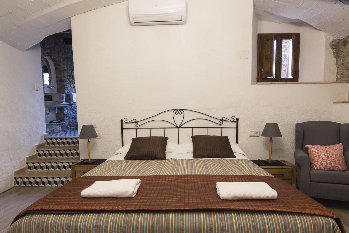 Une chambre à coucher comprenant un lit, deux tables de chevet, une chaise, une fenêtre et un escalier menant à un espace en pierre.