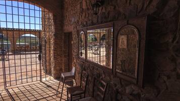 A stone-walled room with a large arched window, a metal gate, and several wooden chairs.