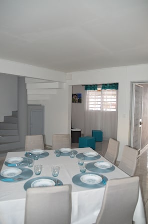 Un coin repas avec une table dressée pour six personnes, des chaises, un escalier et une porte.