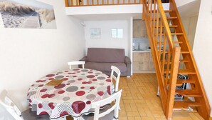 A dining area with a round table, white chairs, and a patterned tablecloth. There is a wooden staircase to the right and a kitchen area in the background. - Cozy Vacation Home Near Beach with Private Garden and Parking (La Tranche-sur-Mer)