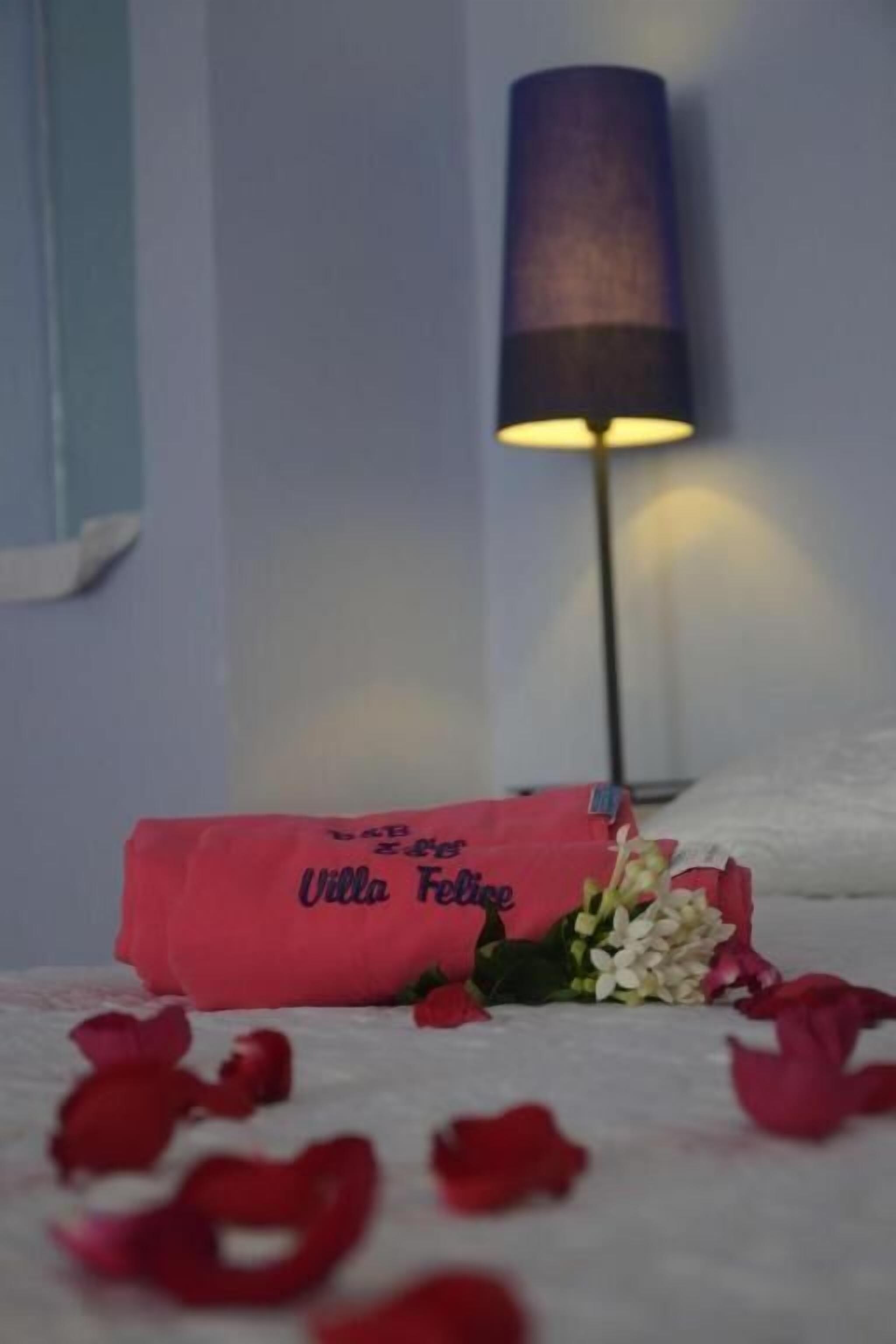 Un bouquet de fleurs, accompagné d’une étiquette portant l’inscription « Villa Felice », est posé sur un lit parsemé de pétales de rose.
