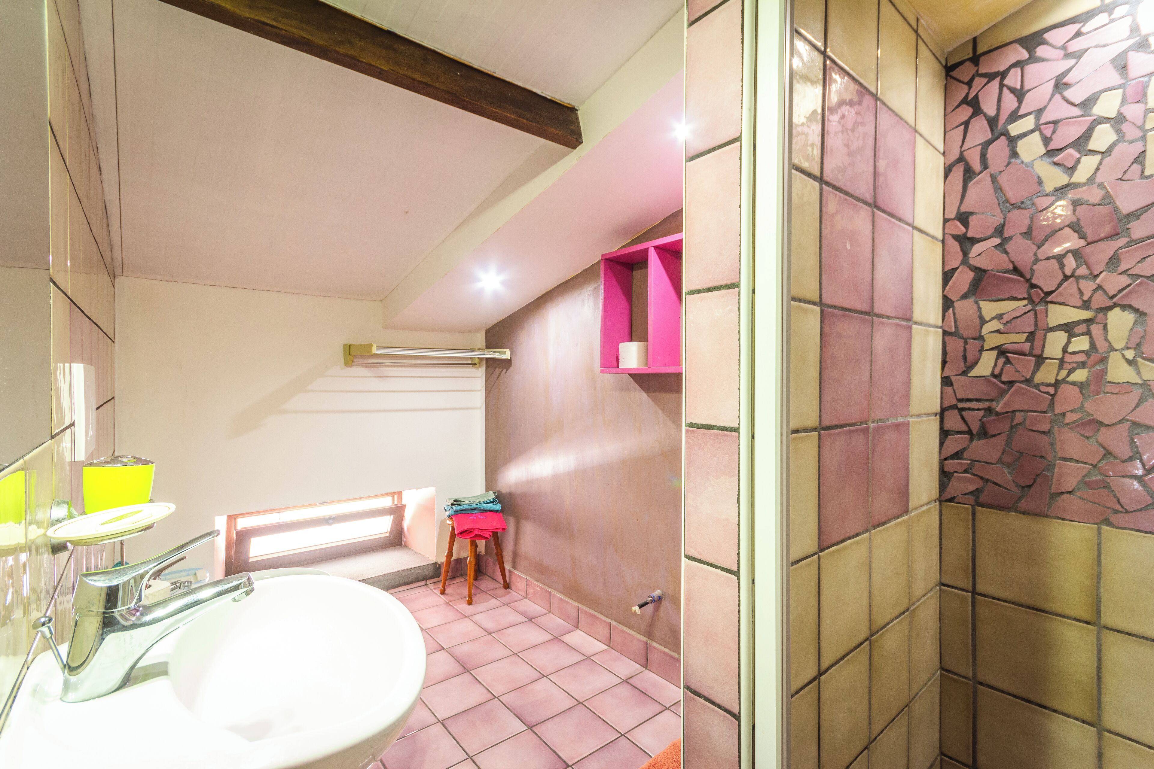 Une salle de bain avec un lavabo blanc, des carreaux roses et beiges, un tabouret en bois et une étagère rose.
