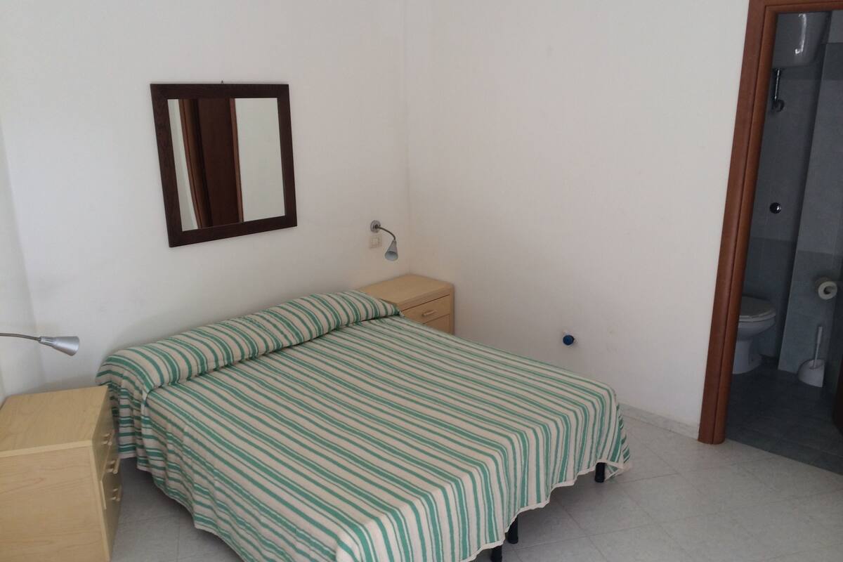 A small, simple bedroom with a single bed, a nightstand, a mirror, and a door leading to a bathroom.